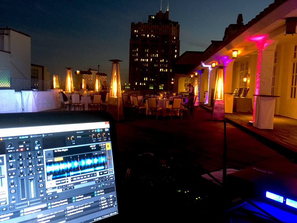 Nighttime rooftop scene with tables, umbrellas, and colorful lighting, and a laptop displaying DJ software in the foreground.