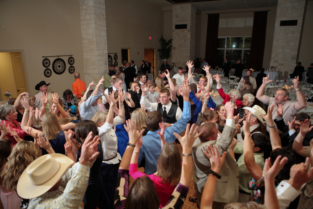 Crowd of people dancing with hands raised at an indoor event, some wearing hats and colorful clothing.