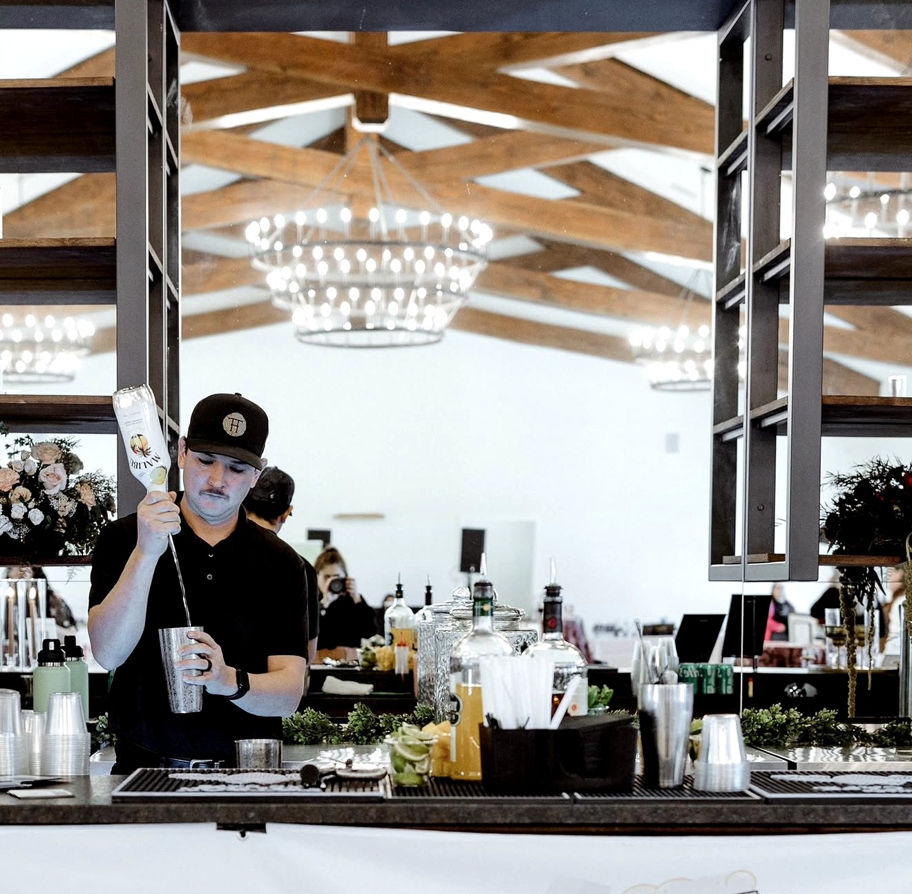Bartender preparing drinks at a bar with a chandelier and wooden ceiling beams in the background.