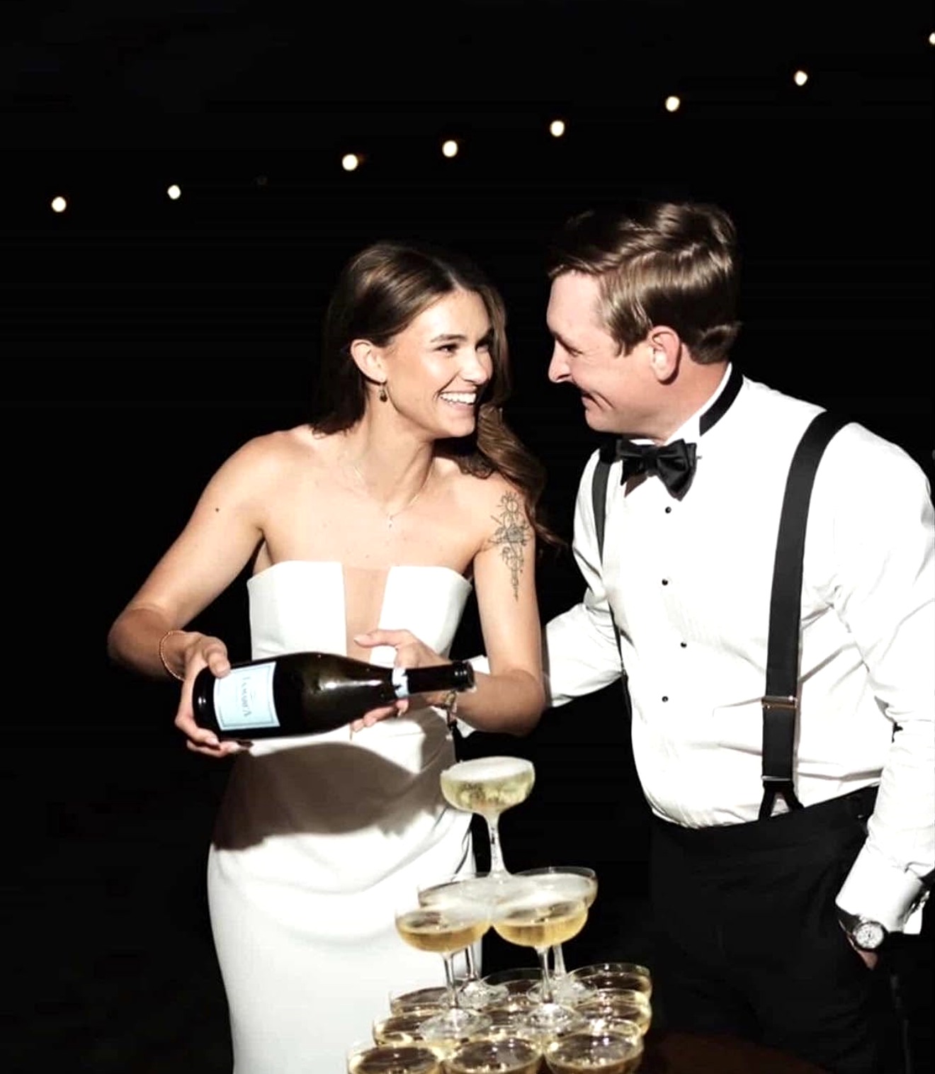 A woman in a white dress and a man in a tuxedo smile and toast with champagne glasses, against a dark background with string lights.