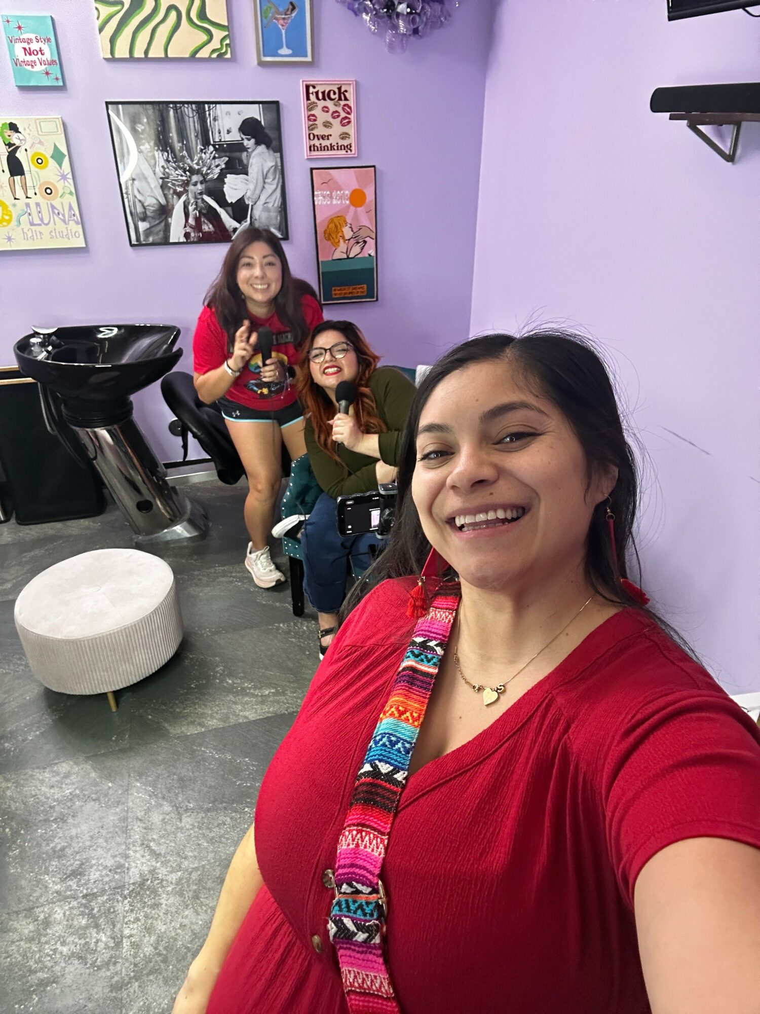 Three women in a purple room, one taking a selfie and two sitting behind, smiling and posing for the photo.