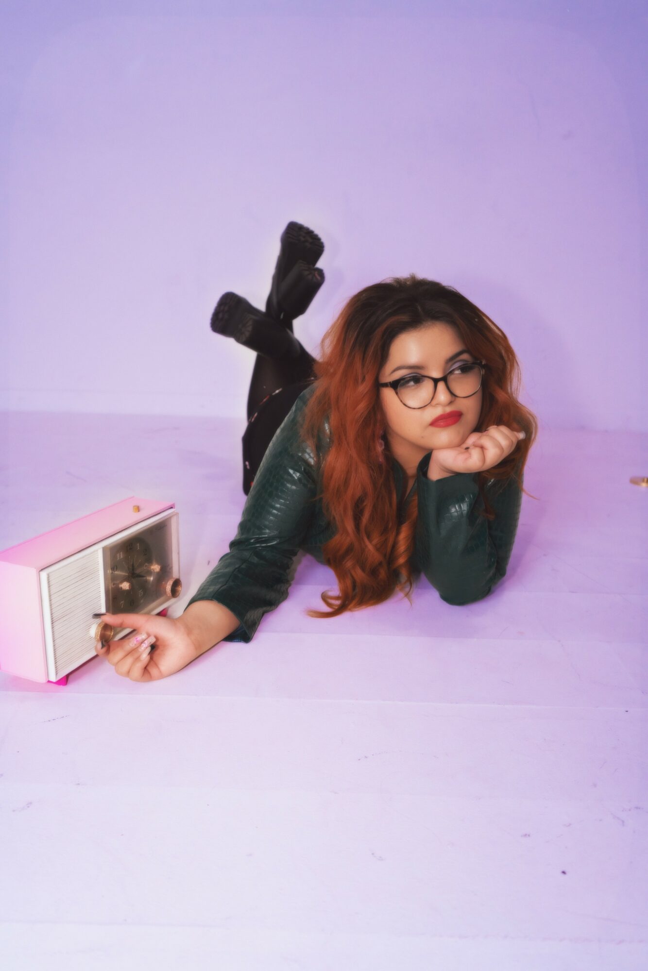 Woman with glasses and curly hair lying on floor, resting her chin on hand, next to a pink radio, against a purple background.