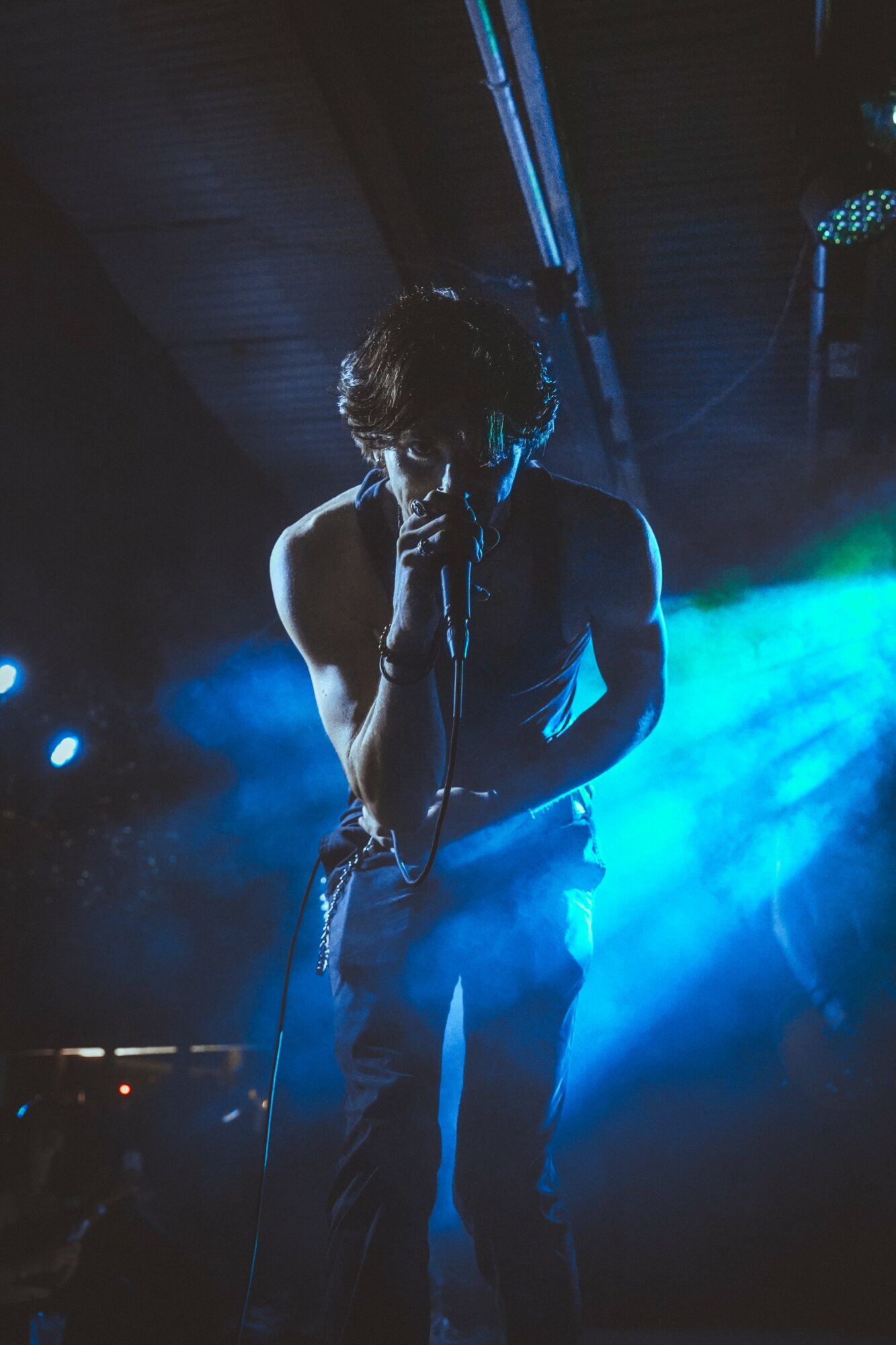 Performer singing into microphone on stage with blue and green lighting, dark background, and visible ceiling beams.