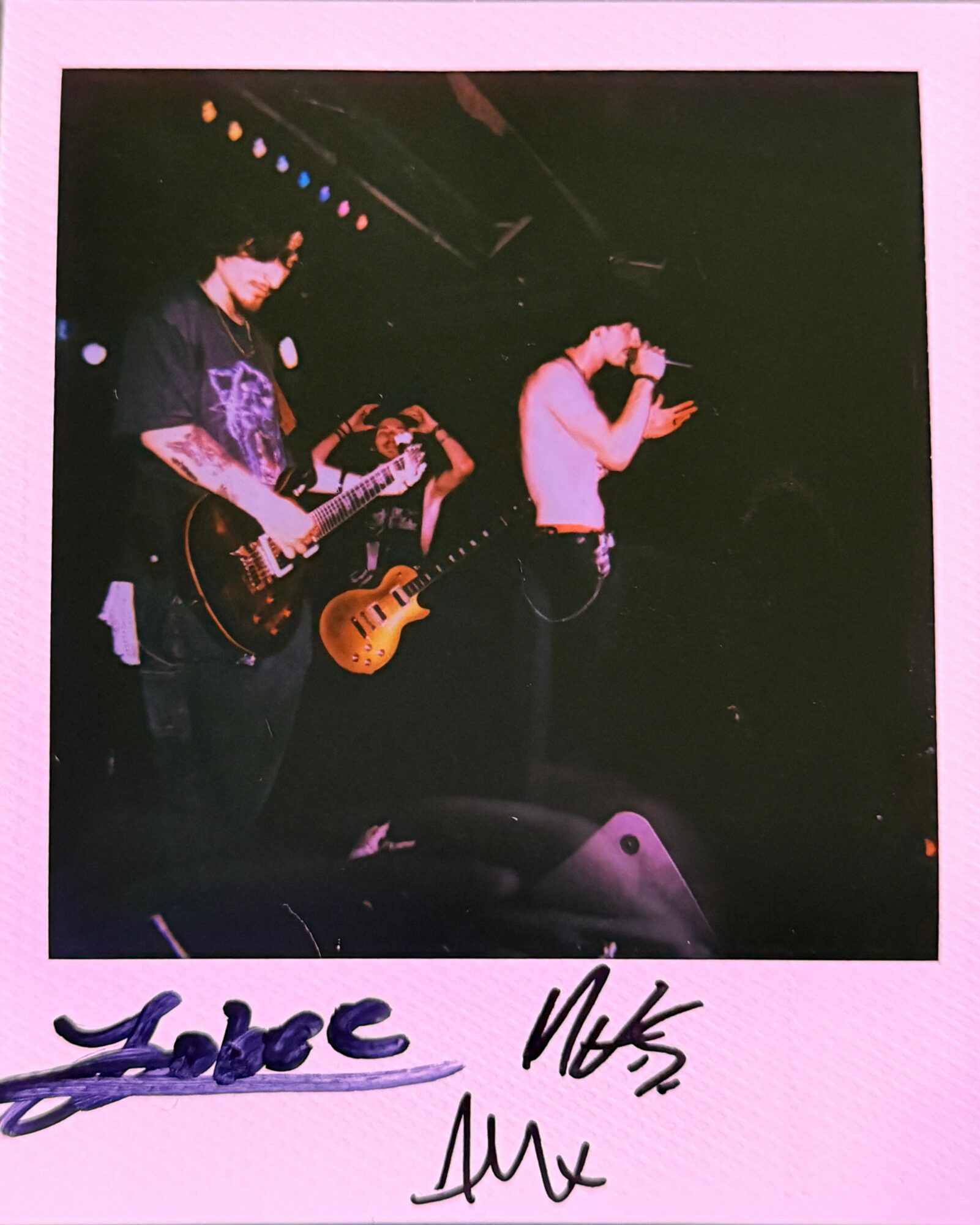 Band performs on stage with two guitarists and a singer, colorful stage lights overhead, pink border, signatures at bottom.