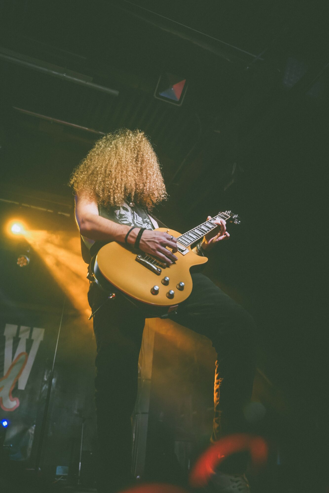 Person with curly hair playing an electric guitar on stage with stage lights in background.
