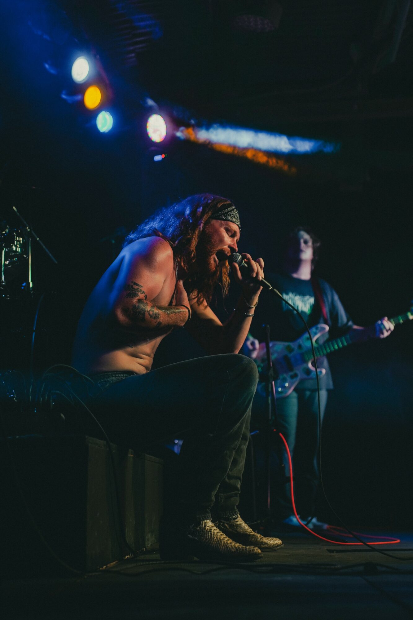 Musician with long hair and tattoos singing into a microphone on stage, with another person playing guitar in the background.