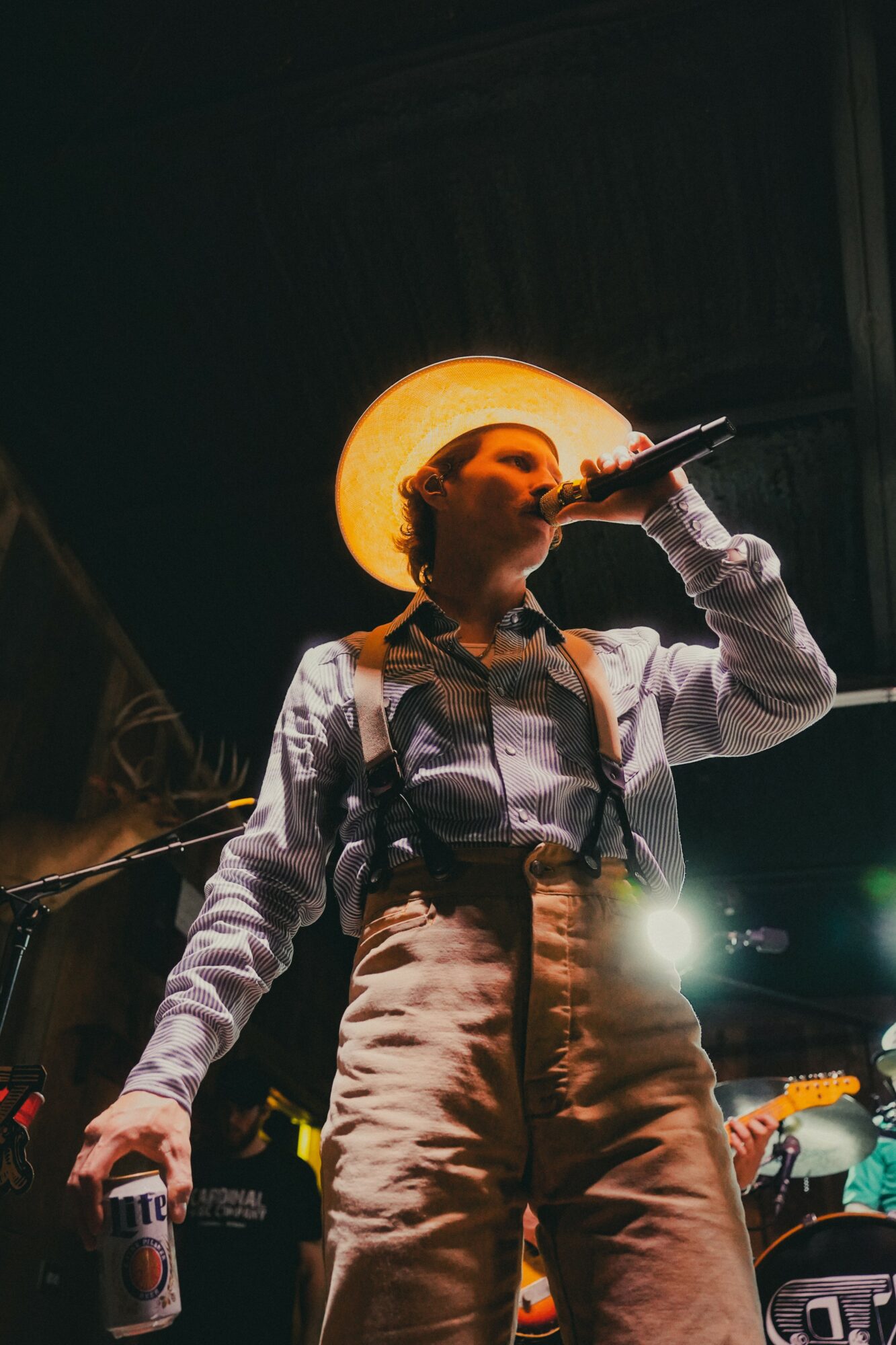 Person singing into microphone on stage with bright light behind, wearing suspenders and high-waisted pants, dark background.
