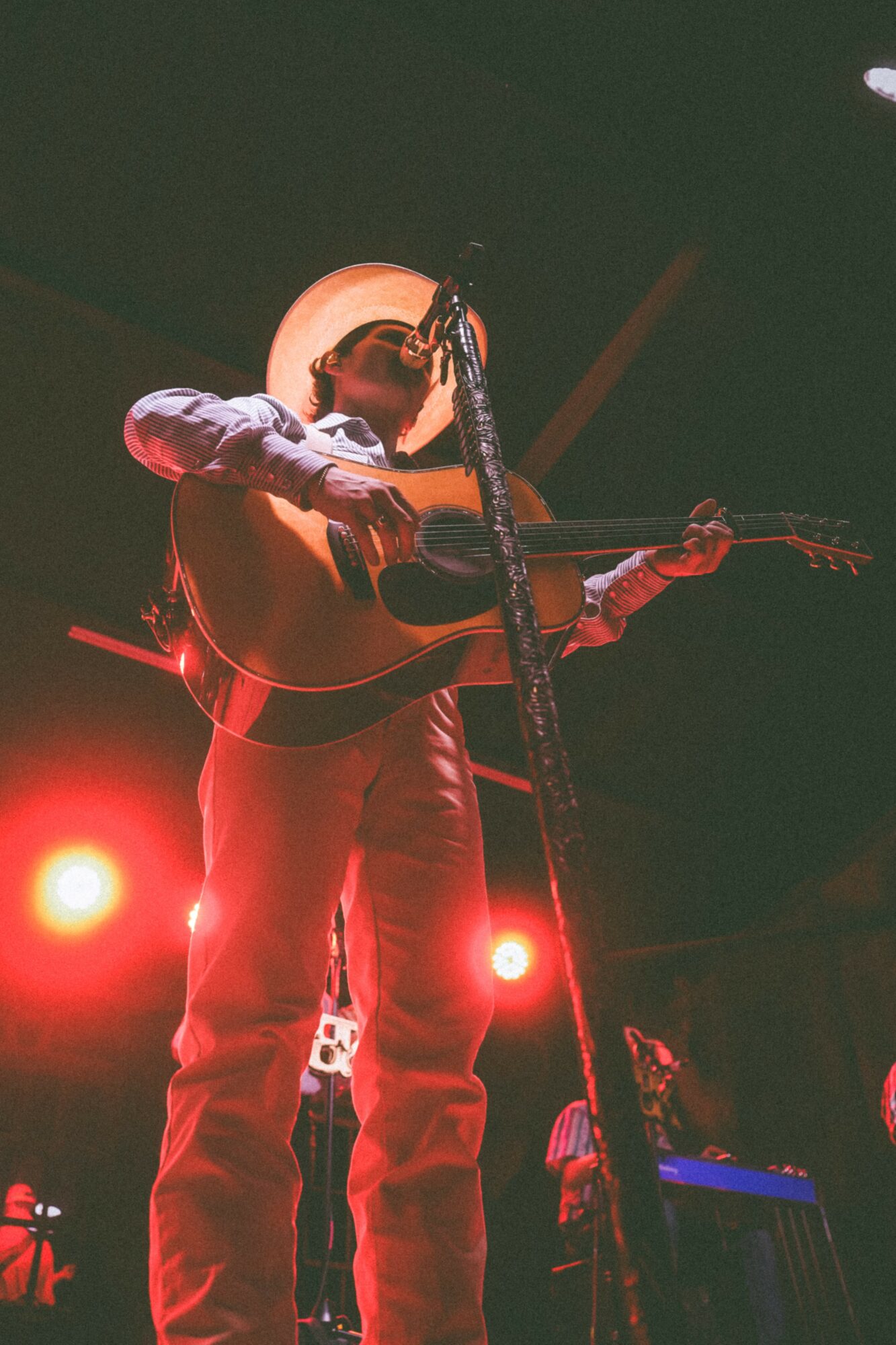 Person wearing a hat playing an acoustic guitar on stage with red lighting and a microphone, facing left.
