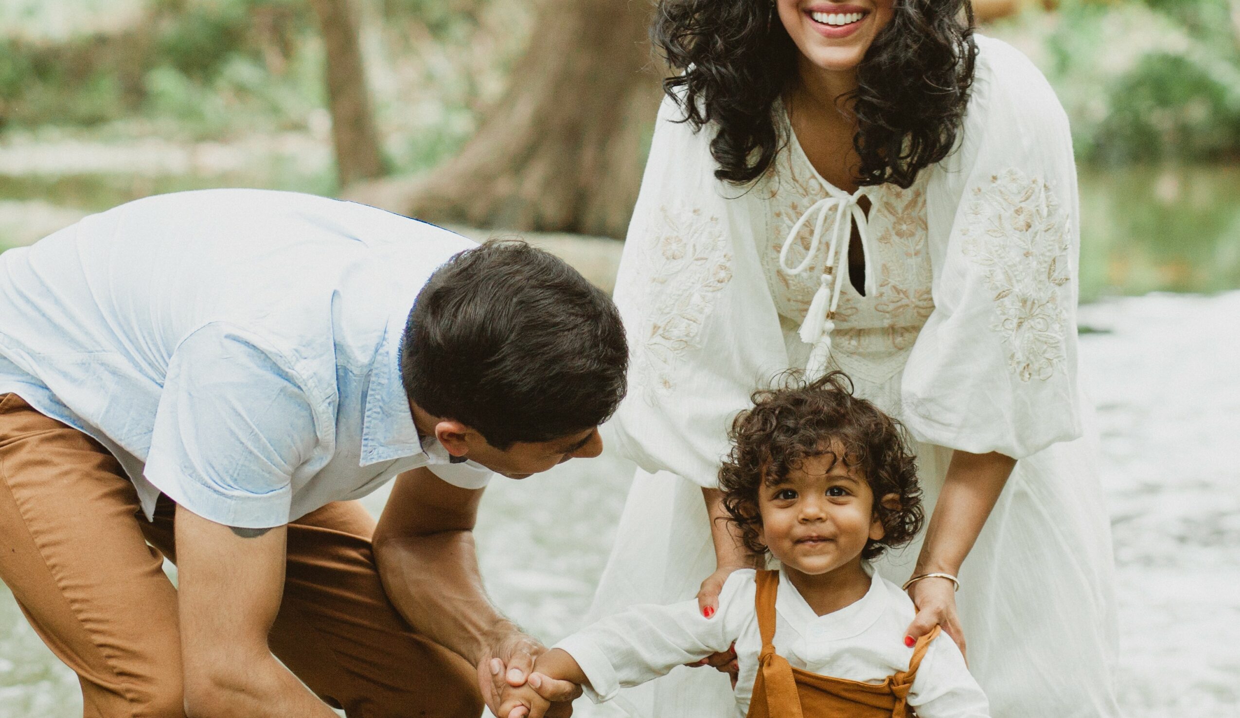 A woman and two children outdoors, smiling and playing on grass, with trees in the background.