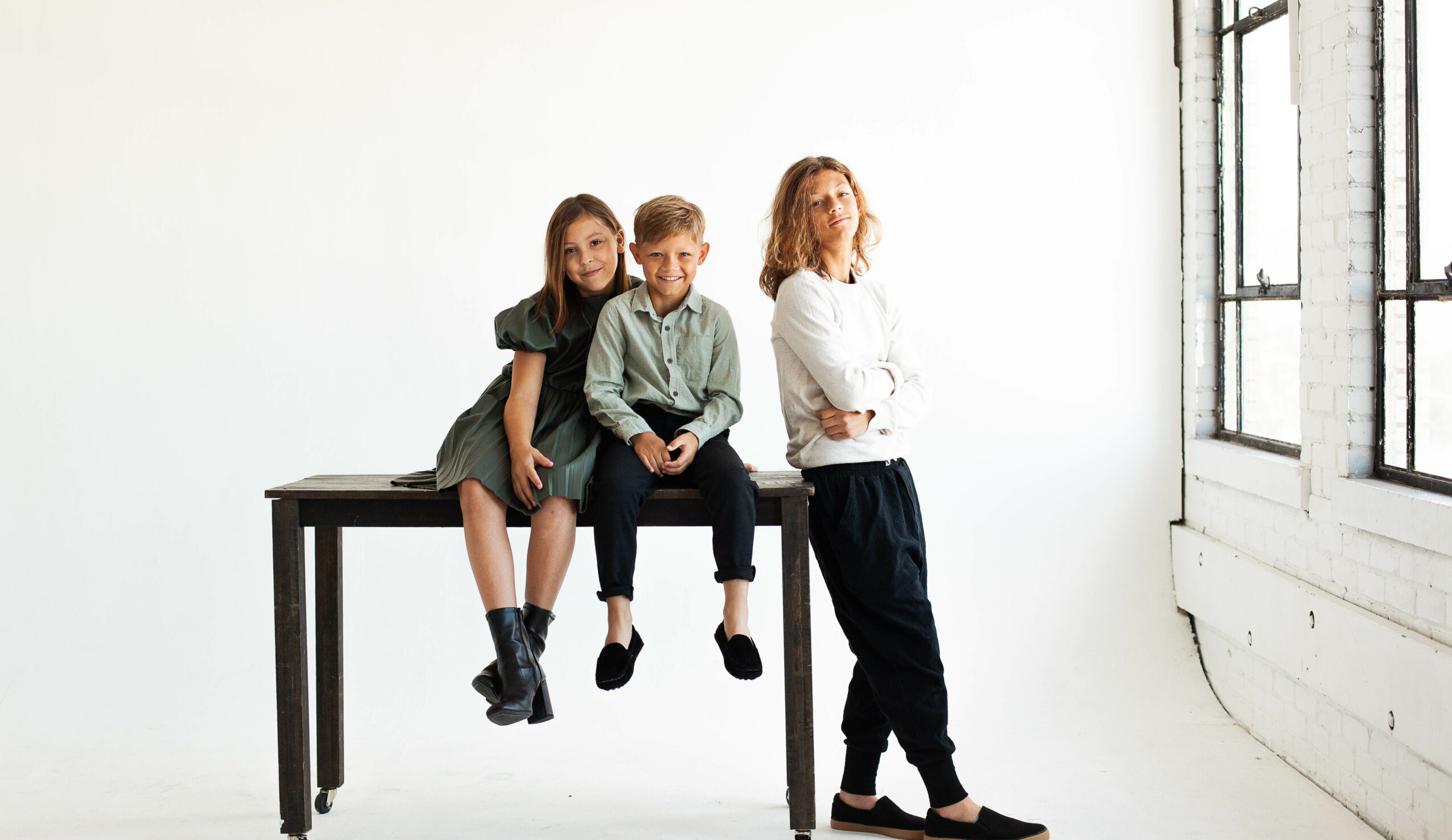 Three children in casual clothing pose near a window in a bright room, with one girl sitting on a table and another standing.