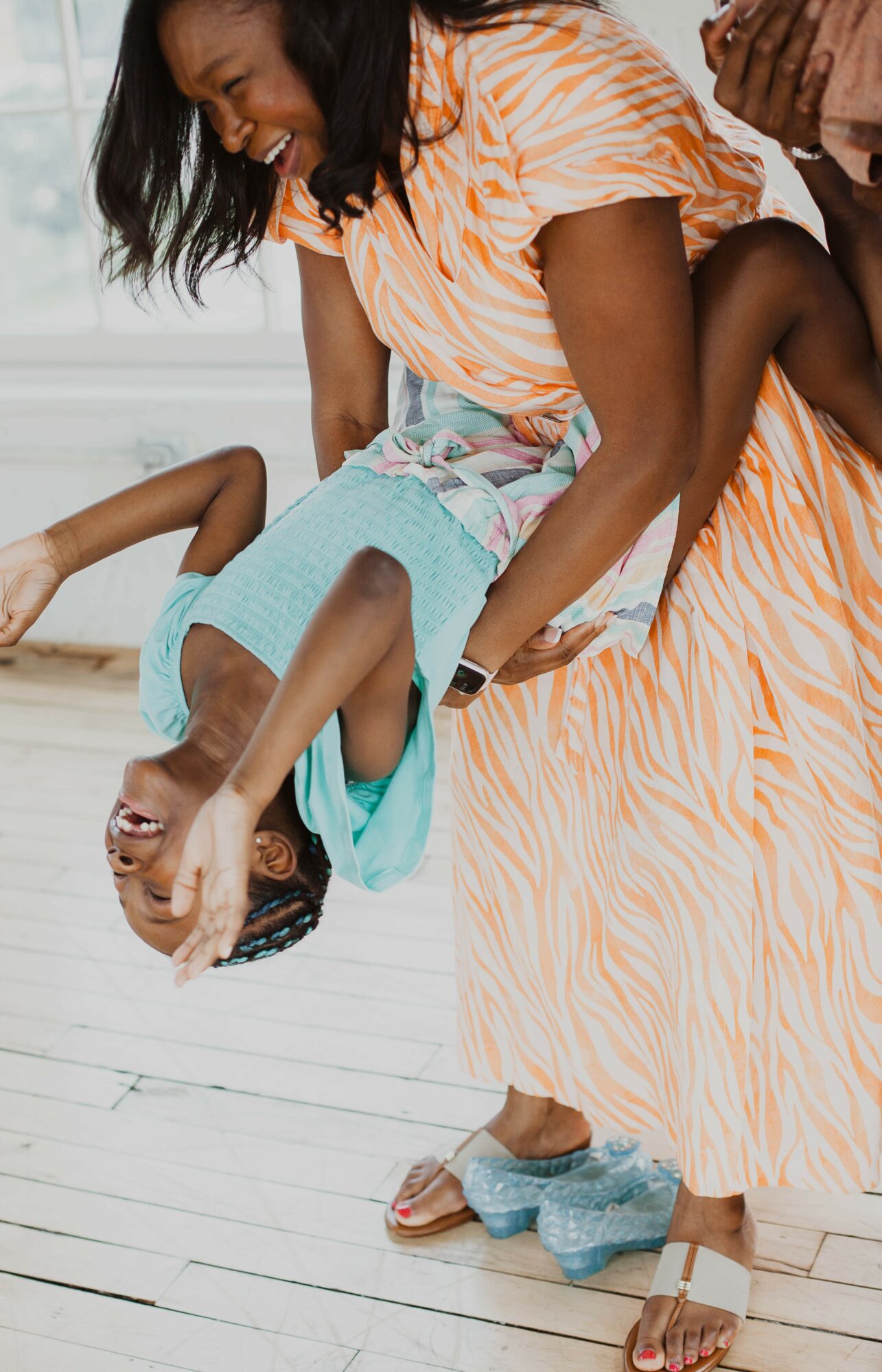 Two women and a child playing indoors, woman holding child upside down, smiling, child wearing glasses, wooden floor background.