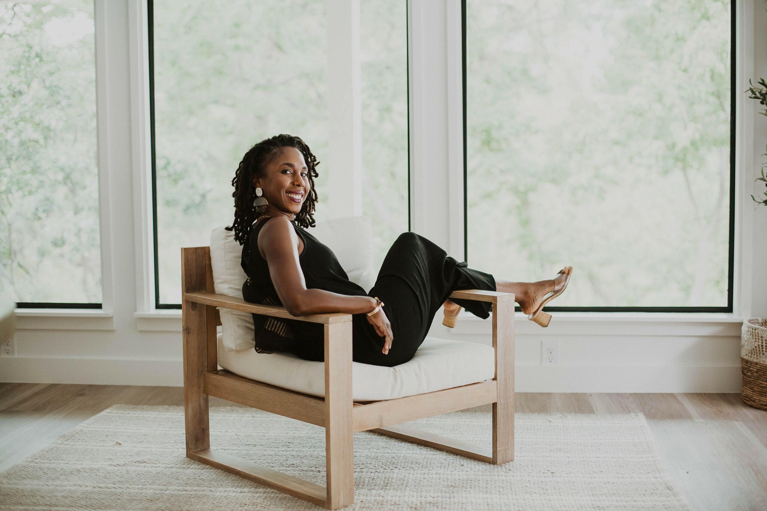 Woman sitting on a wooden chair in front of large windows, with a neutral expression and relaxed pose.