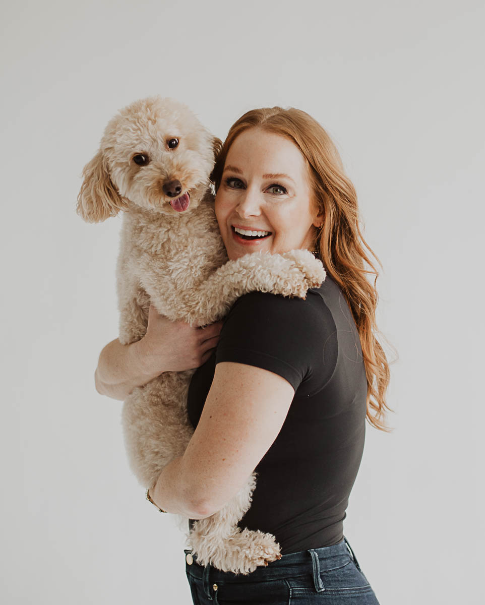 Woman with red hair smiling and holding a small, curly-haired dog against a plain background.
