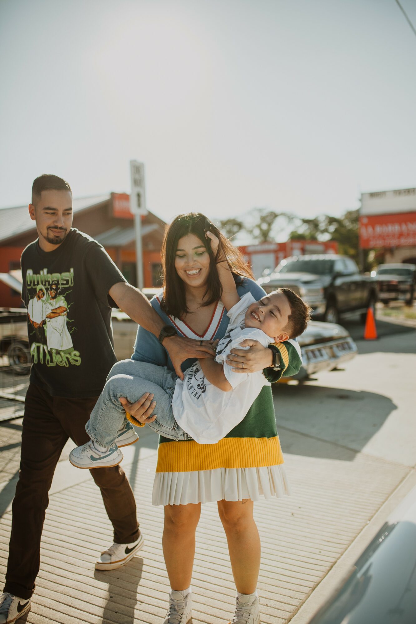 A woman carrying a child while smiling, with a man walking beside them outdoors in a parking lot.