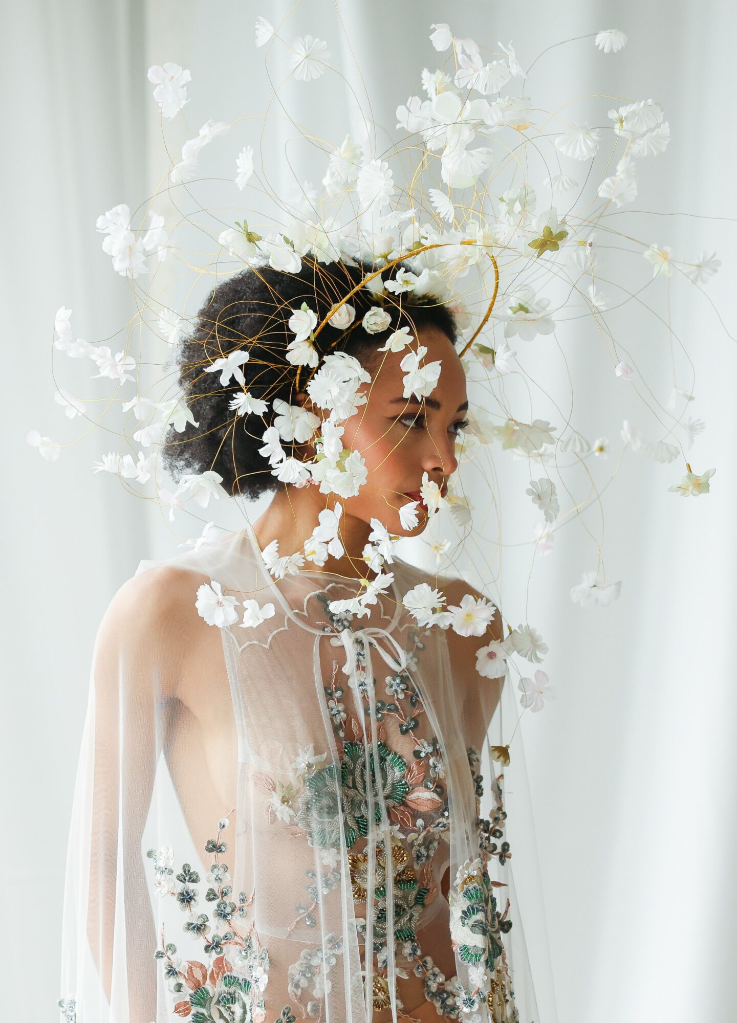 Woman with floral headpiece and sheer dress, facing right, standing in front of white curtains.