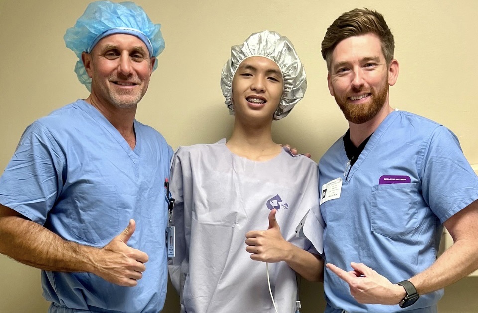 Three medical professionals in scrubs and caps smiling and giving thumbs up.