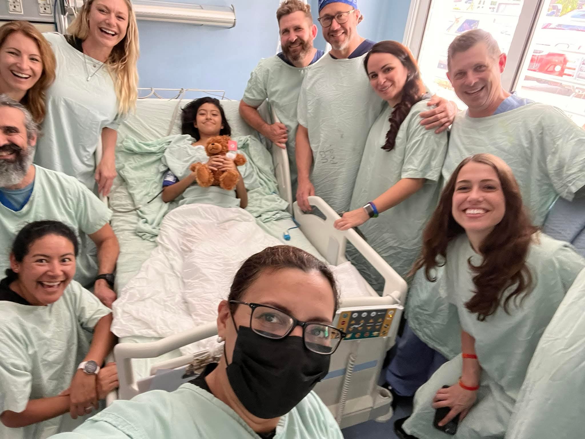 Group of people in hospital gowns gathered around a patient in bed, smiling and celebrating, with a nurse in the foreground wearing a mask.
