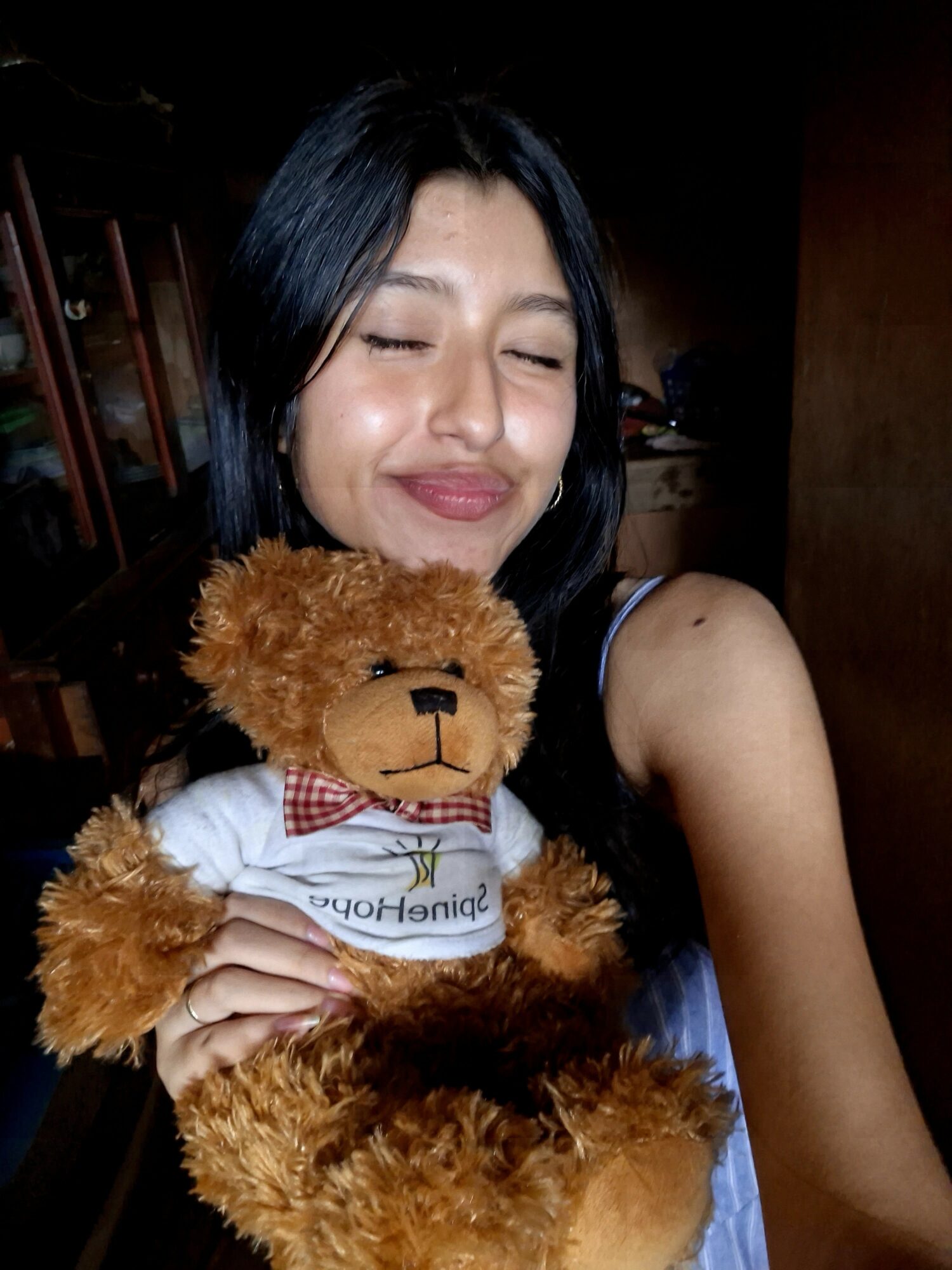 Young woman with black hair holding a teddy bear, smiling with eyes closed, in a dimly lit room.