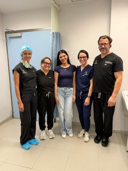 Five healthcare professionals standing in a hospital room, smiling, with a curtain and door in the background.