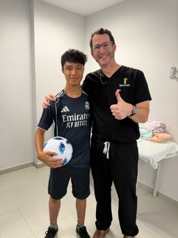 Two people standing in a room, one holding a soccer ball, smiling, with a bed in the background.