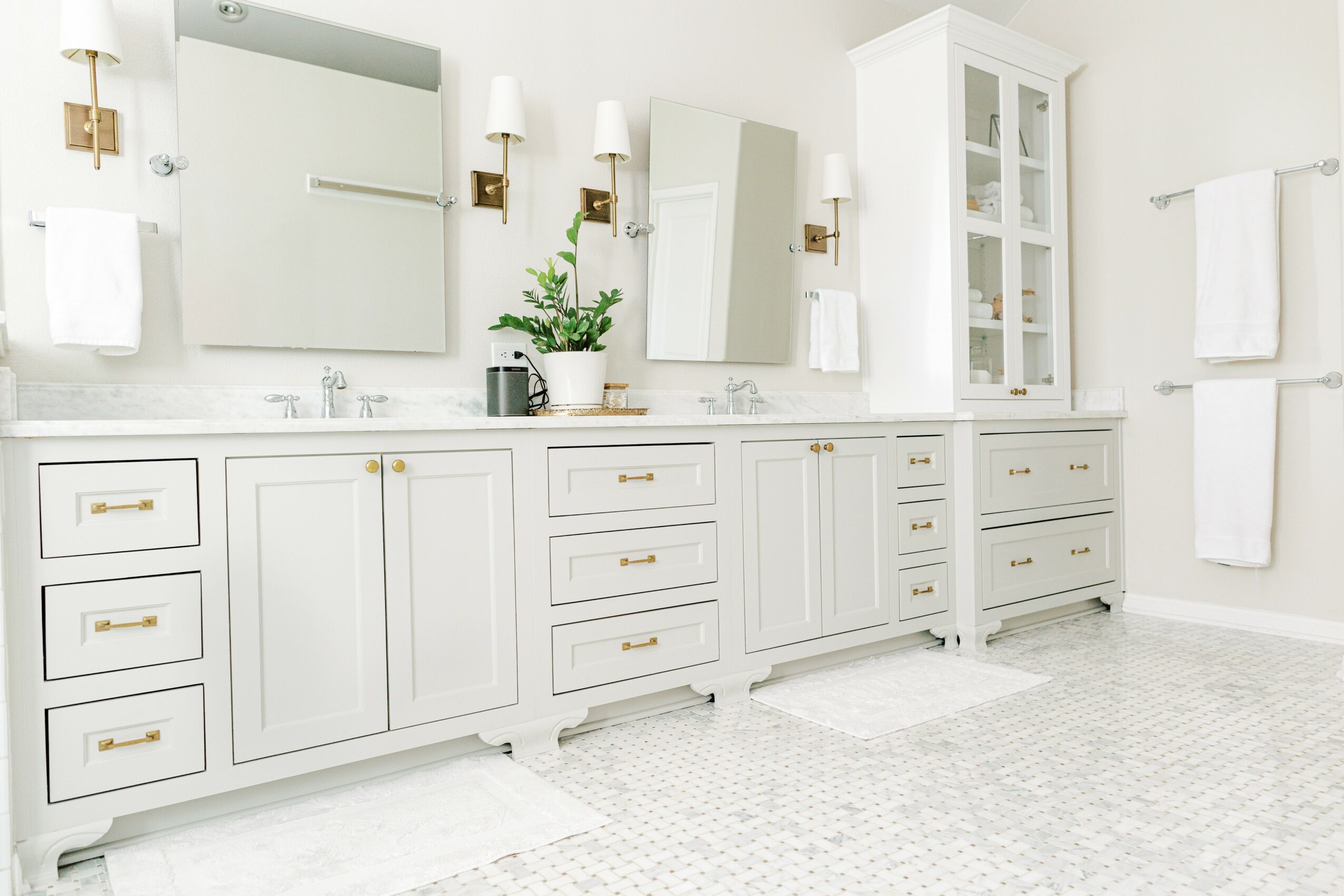 Bright bathroom with white cabinets, mirror, towels, and a plant on the countertop, illuminated by wall-mounted lights.