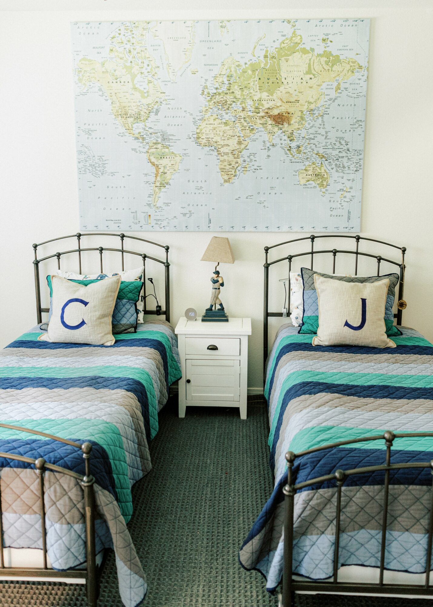 Two beds with striped blue, green, and white bedding, pillows with letters, a nightstand with a lamp, and a world map on the wall.
