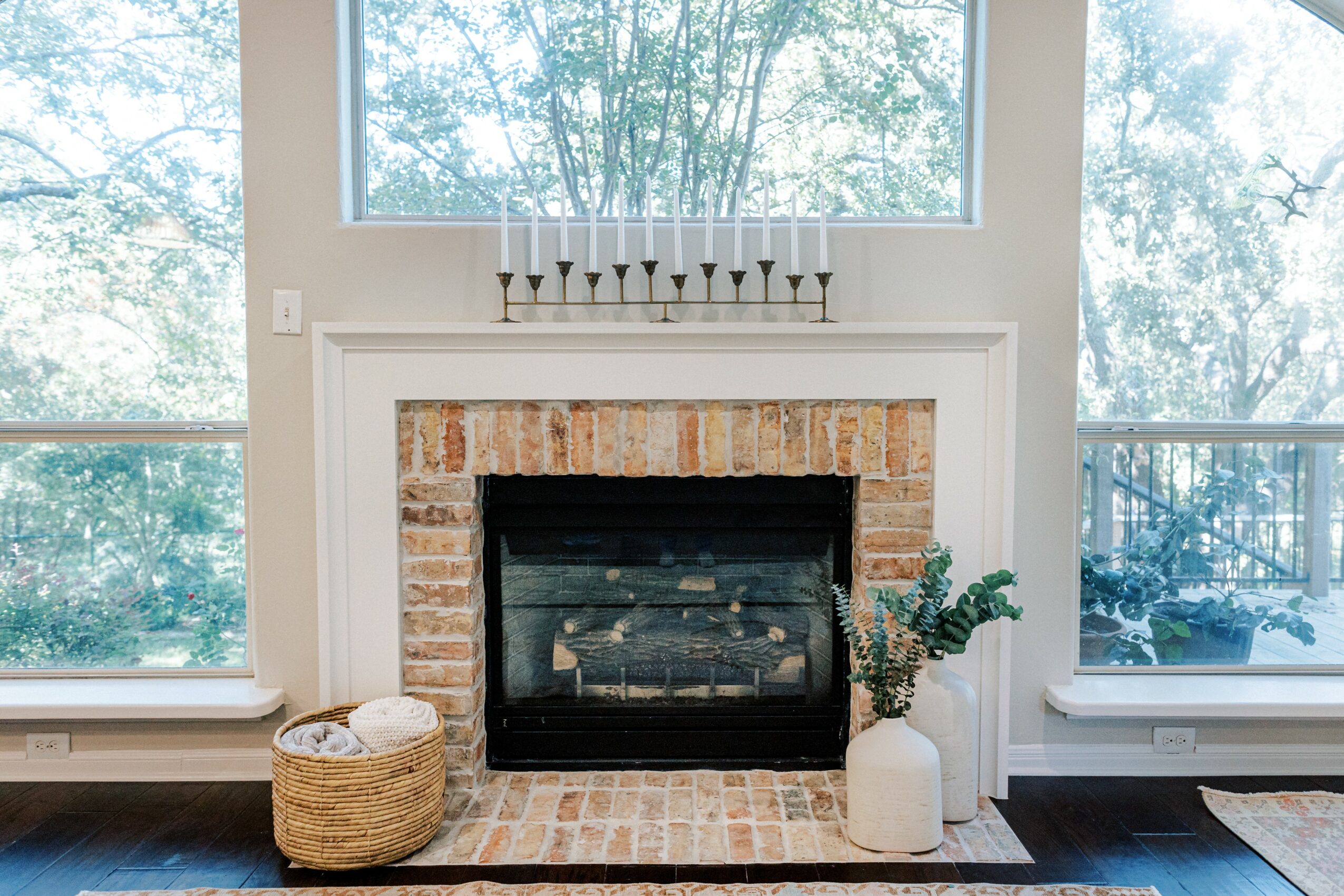 Fireplace with brick surround, white mantel, and a menorah on top, flanked by large windows showing trees outside.