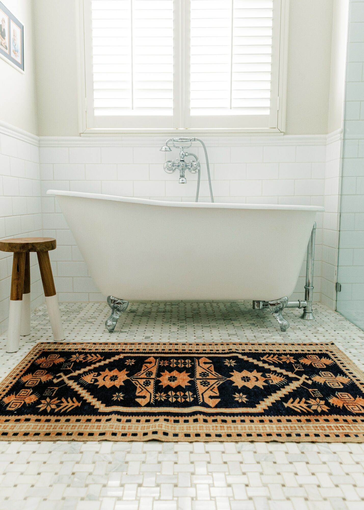 White bathtub with clawfoot legs in a bathroom with white tiled walls and a window with shutters, a wooden stool, and a patterned rug.