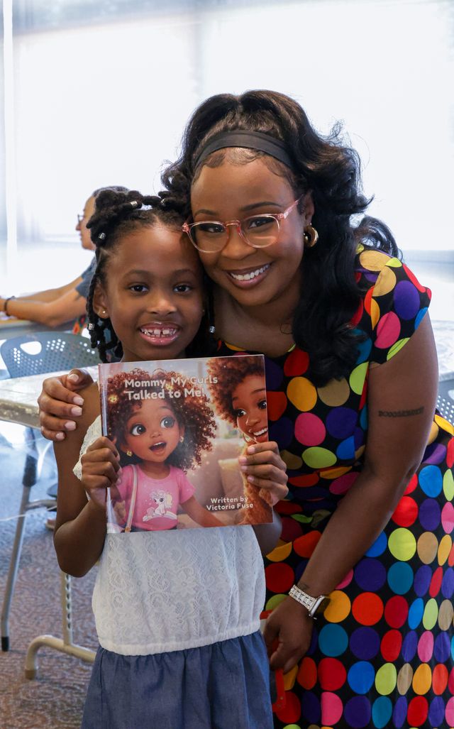 Smiling woman and girl holding a magazine with a cartoon girl on the cover, indoors with bright background.