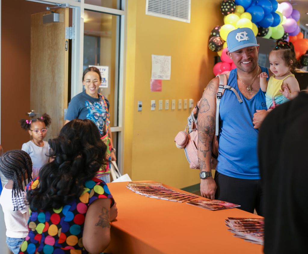 People gather at a colorful indoor event, with balloons and a table, smiling and interacting. Child in yellow held by man.