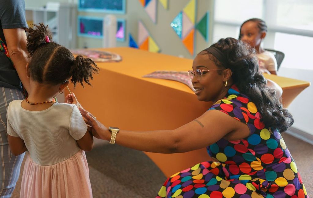 Woman kneeling and holding hands with a young girl in a colorful dress, in a bright room with decorations.