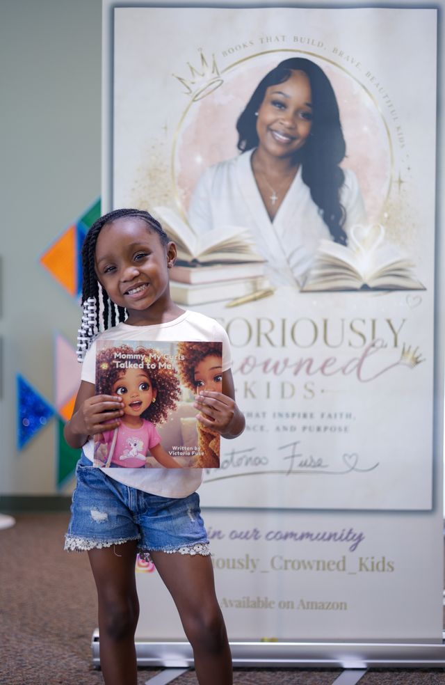 Young girl smiling and holding a children's book in front of a poster with a woman and open books, indoors.