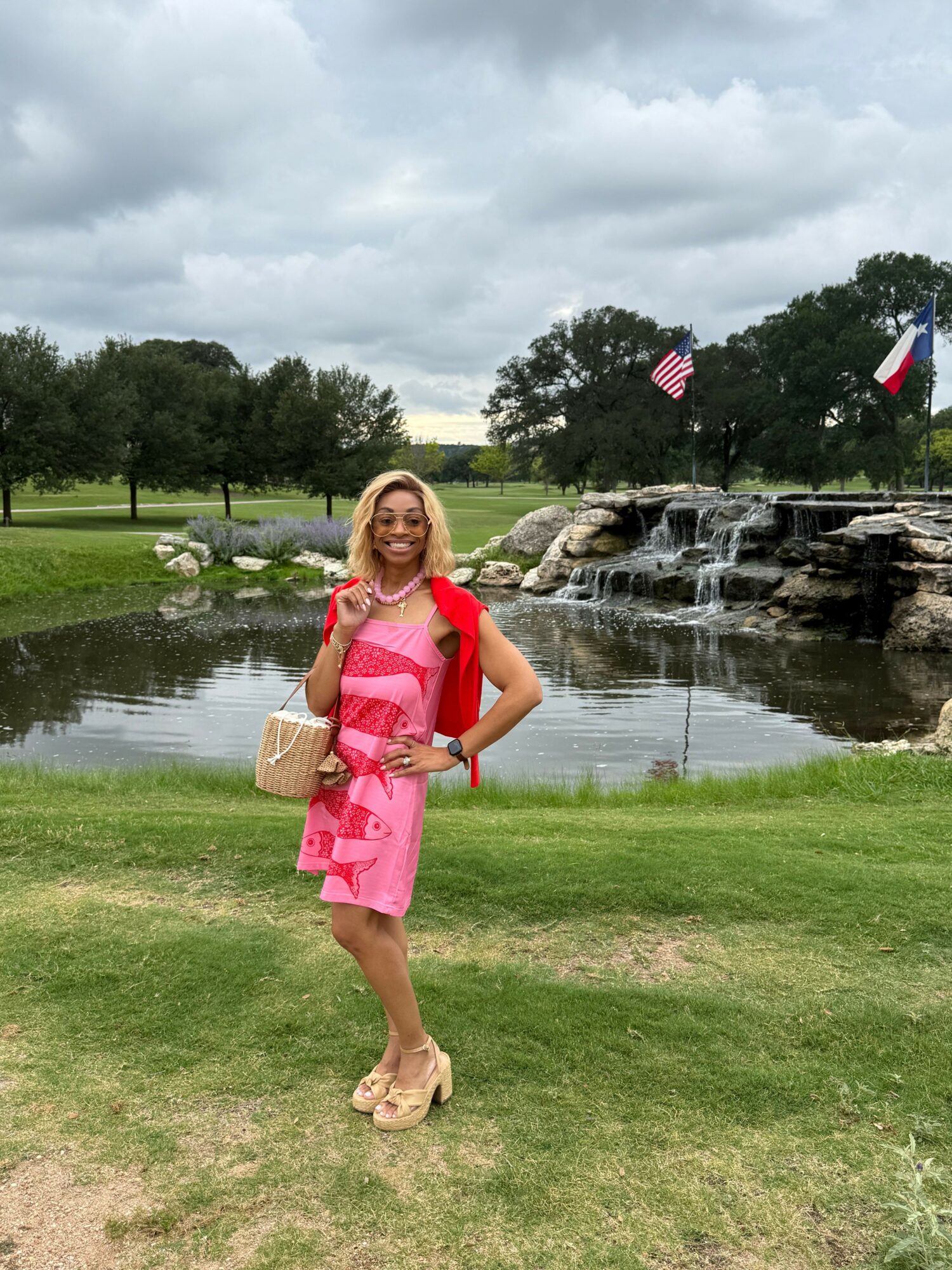 Woman in pink dress and beige shoes standing on grass near a pond with flags and waterfall in background.