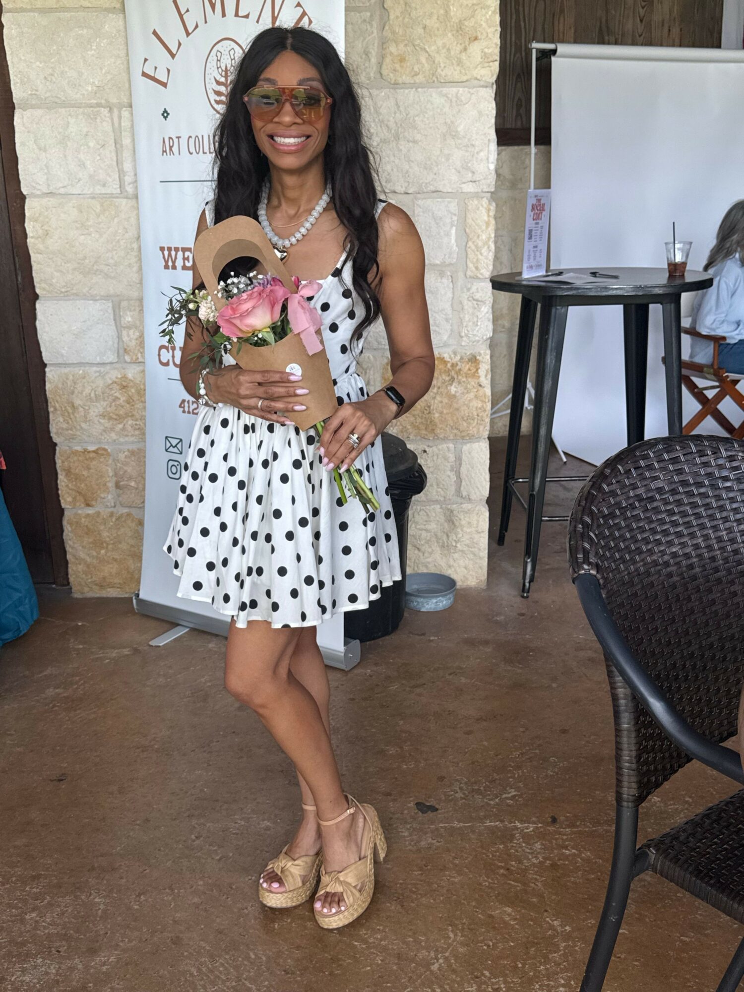 Woman in polka dot dress holding a bouquet of pink roses, smiling, standing indoors with a sign and furniture behind her.