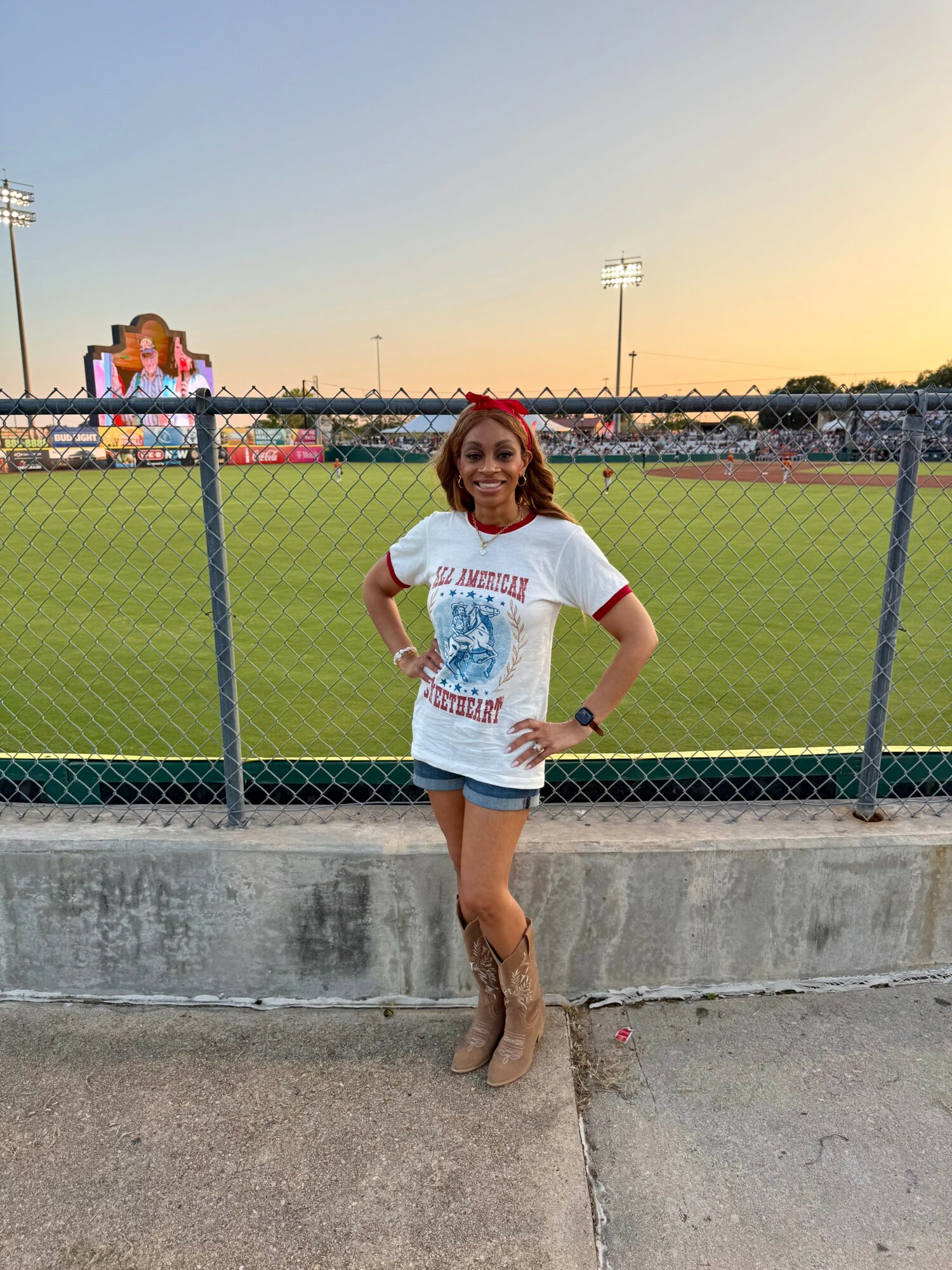 Young woman with red hair standing outdoors near a chain-link fence, smiling, with a sports field and sunset in background.
