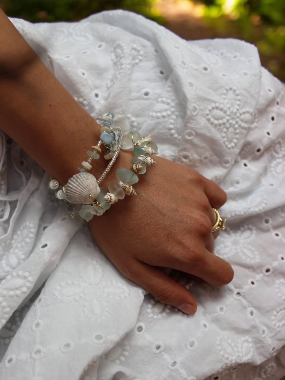 Close-up of a hand resting on white embroidered fabric, wearing multiple bracelets with beads and shells, and a gold ring.