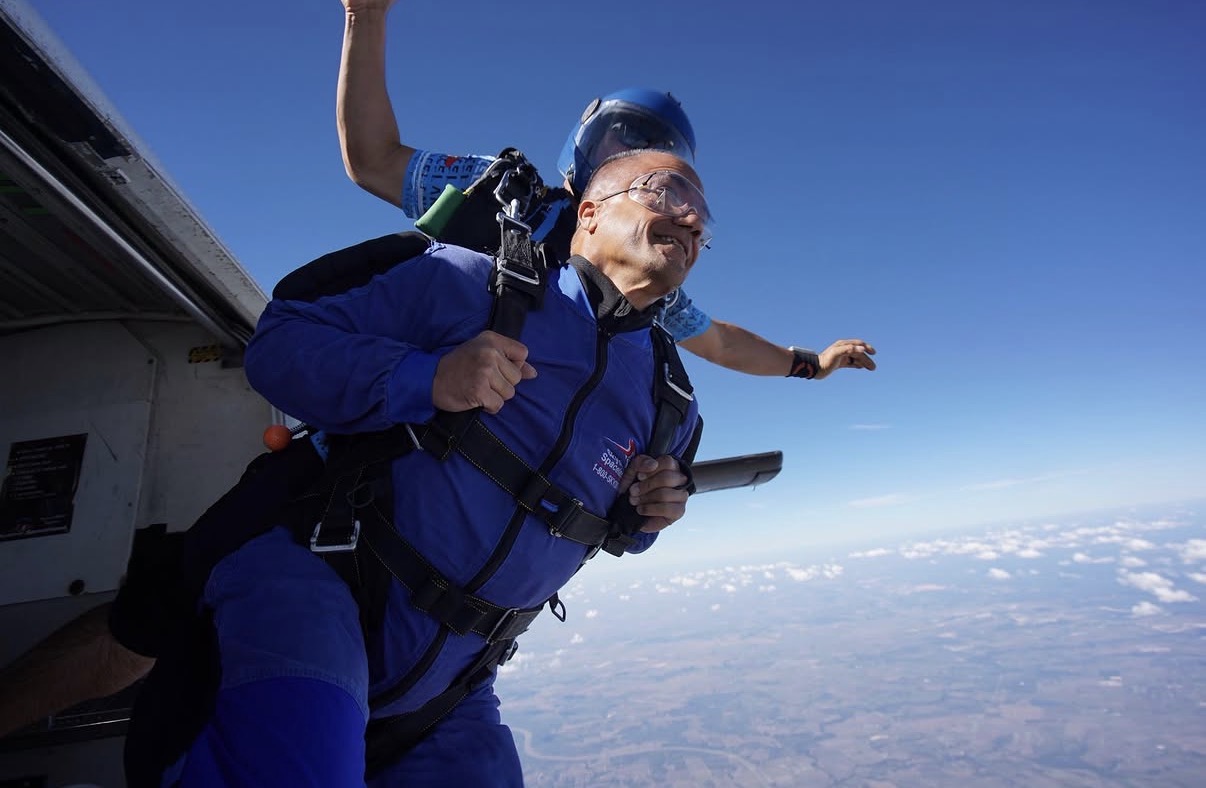 Person smiling and holding a book, sky and clouds in background, skydiving exit scene, clear weather, bright sunlight.