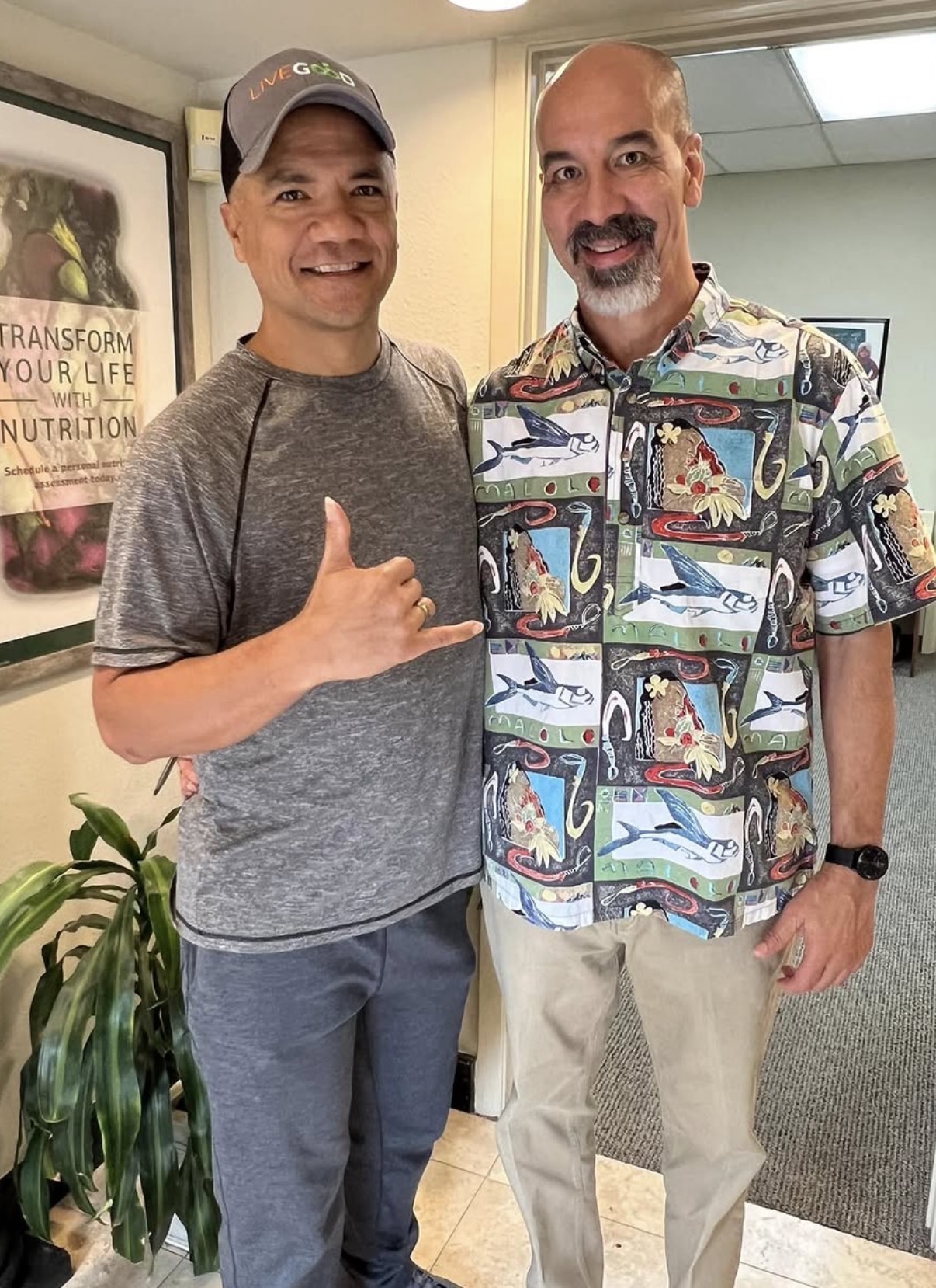 Two men standing indoors, smiling, one making a shaka sign, with a plant and framed poster in the background.