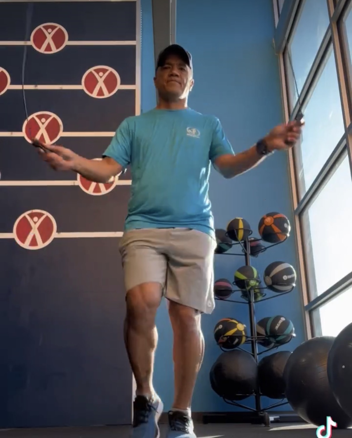 Person balancing on one leg with arms outstretched indoors, sports equipment nearby, sunlight through windows.
