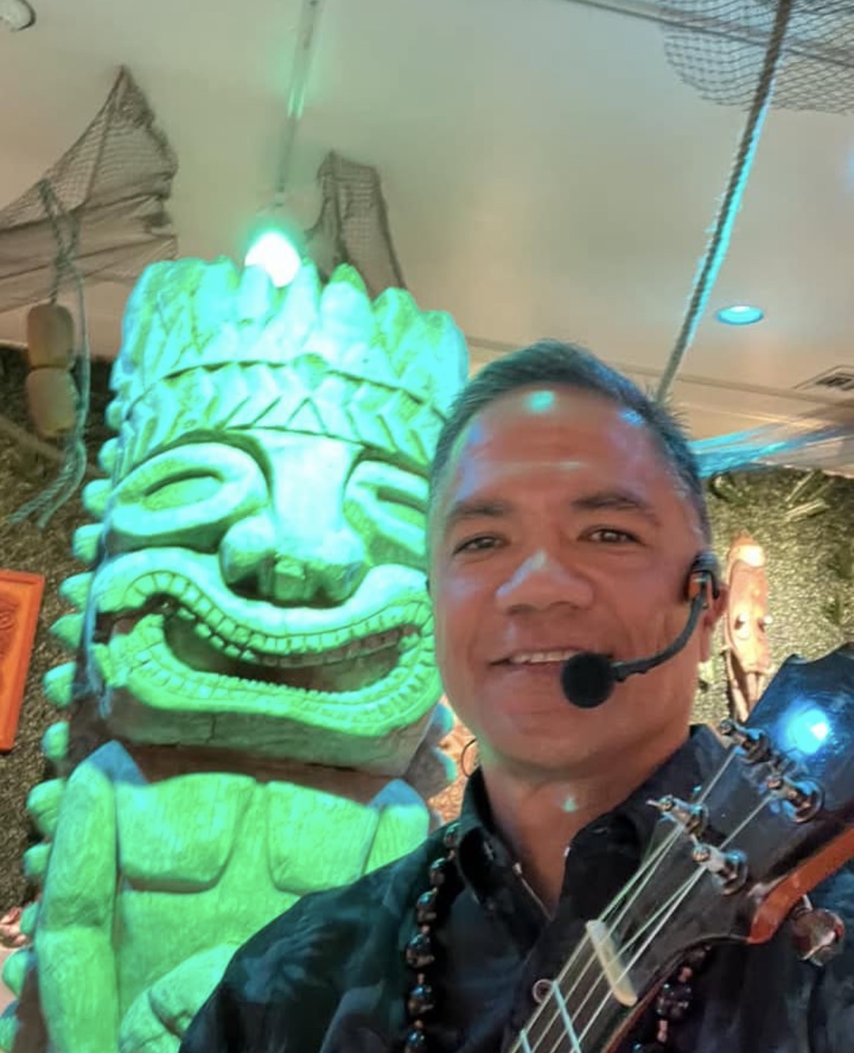 Man wearing a headset microphone smiling in front of a large, glowing green tiki statue indoors.