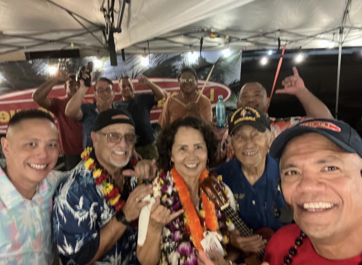 Group of people smiling and celebrating under a tent at night, some wearing leis and hats.