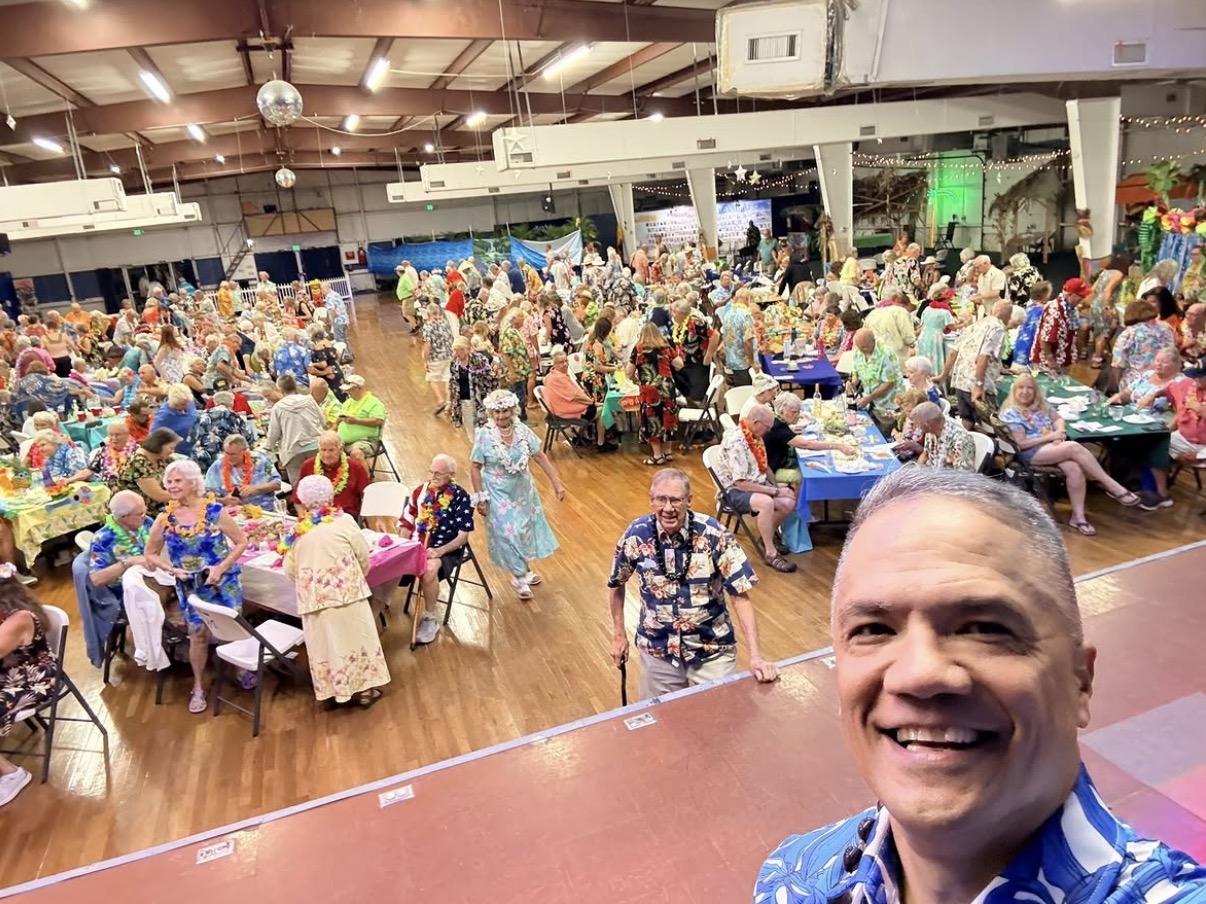 A man smiling in the foreground with a large crowded indoor event in the background, filled with people at tables and decorations.