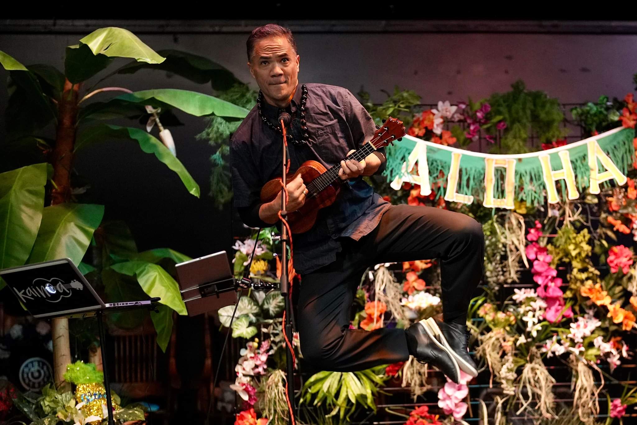 Man playing ukulele on stage with tropical plants and colorful flowers, ALOHA banner in background.