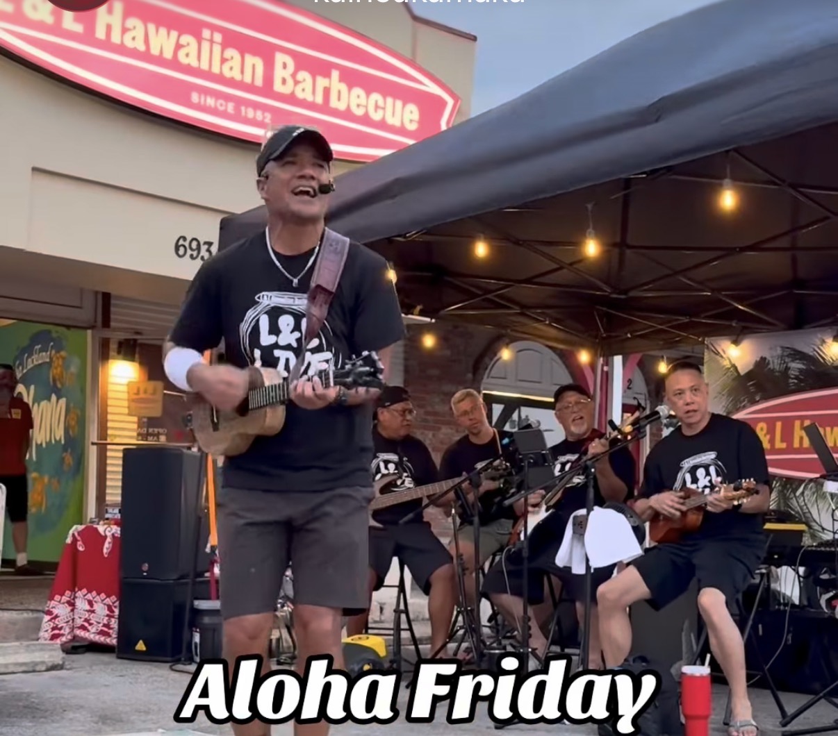 Group of musicians performing outdoors under a tent, with a sign for Hawaiian Barbecue in the background, and a person singing and playing ukulele.