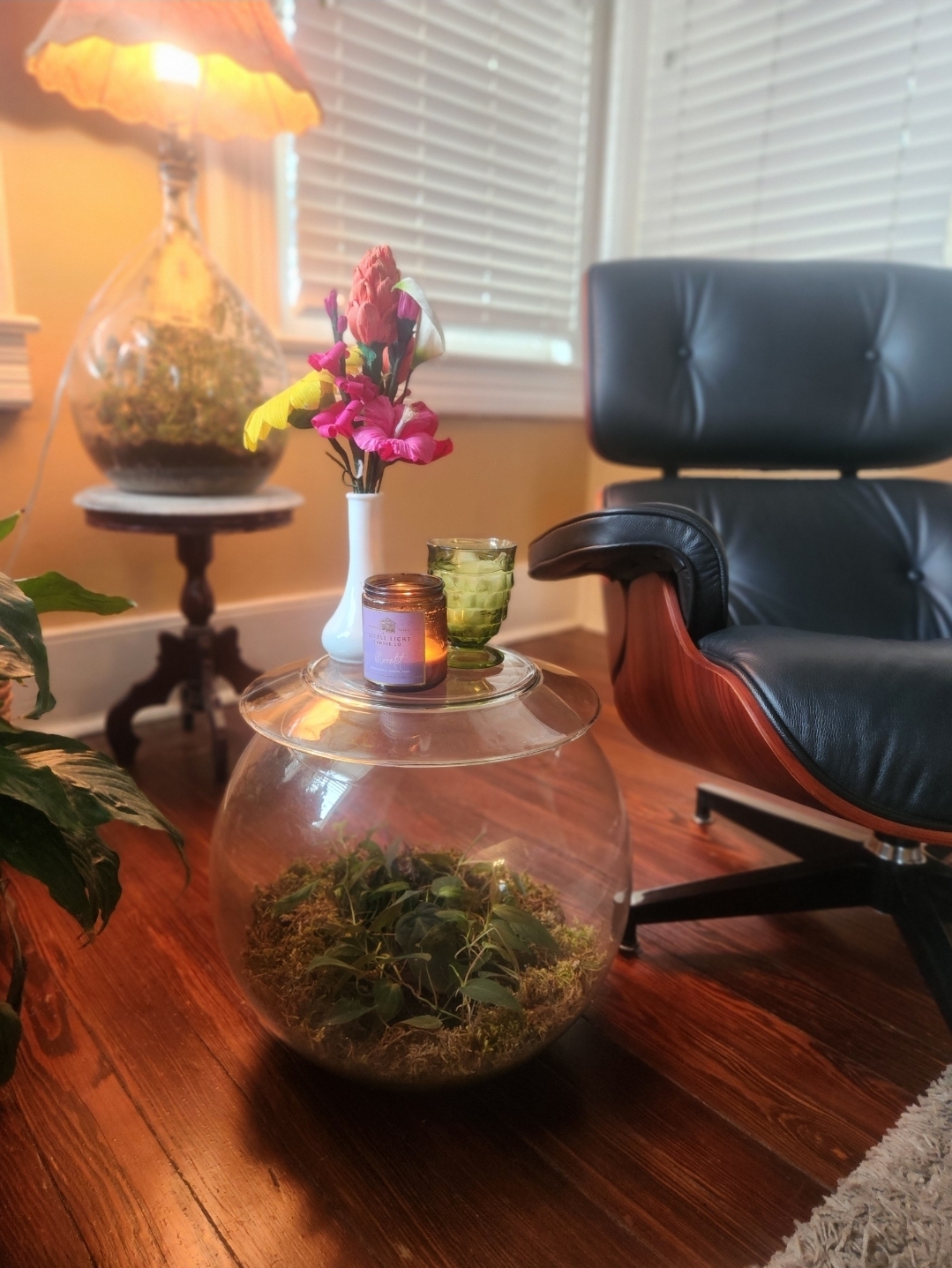 Glass bowl with plants on wooden table, pink flowers in a white vase, lamp, and black leather chair in background.