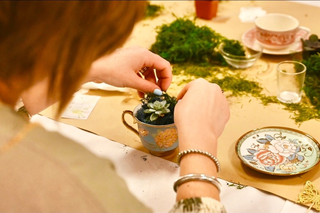 Person decorating a small teapot with a plant, surrounded by dishes and greenery on a table.