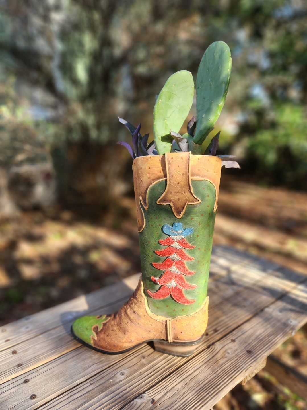 Decorative cowboy boot planter with green plants, placed on a wooden surface outdoors, background with blurred trees and sunlight.