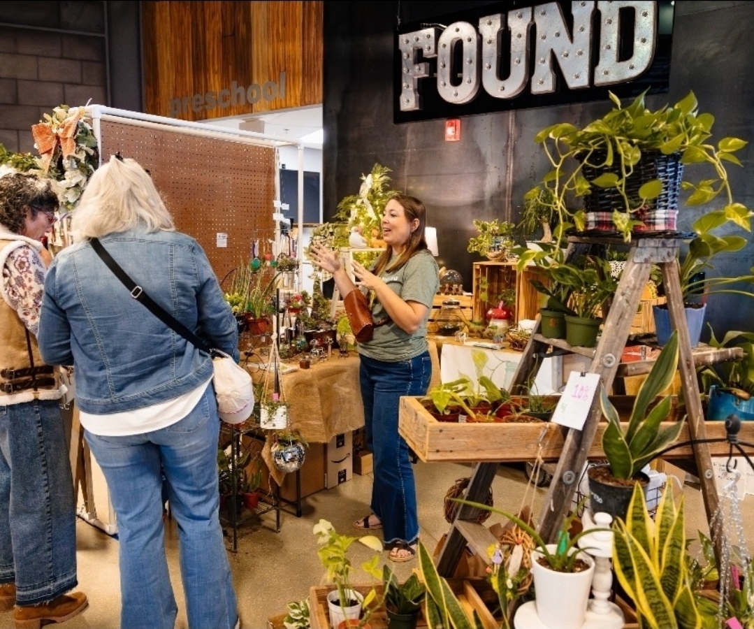 People browsing a plant and flower shop with a woman taking a photo, plants on shelves, and a large 'FOUND' sign on wall.