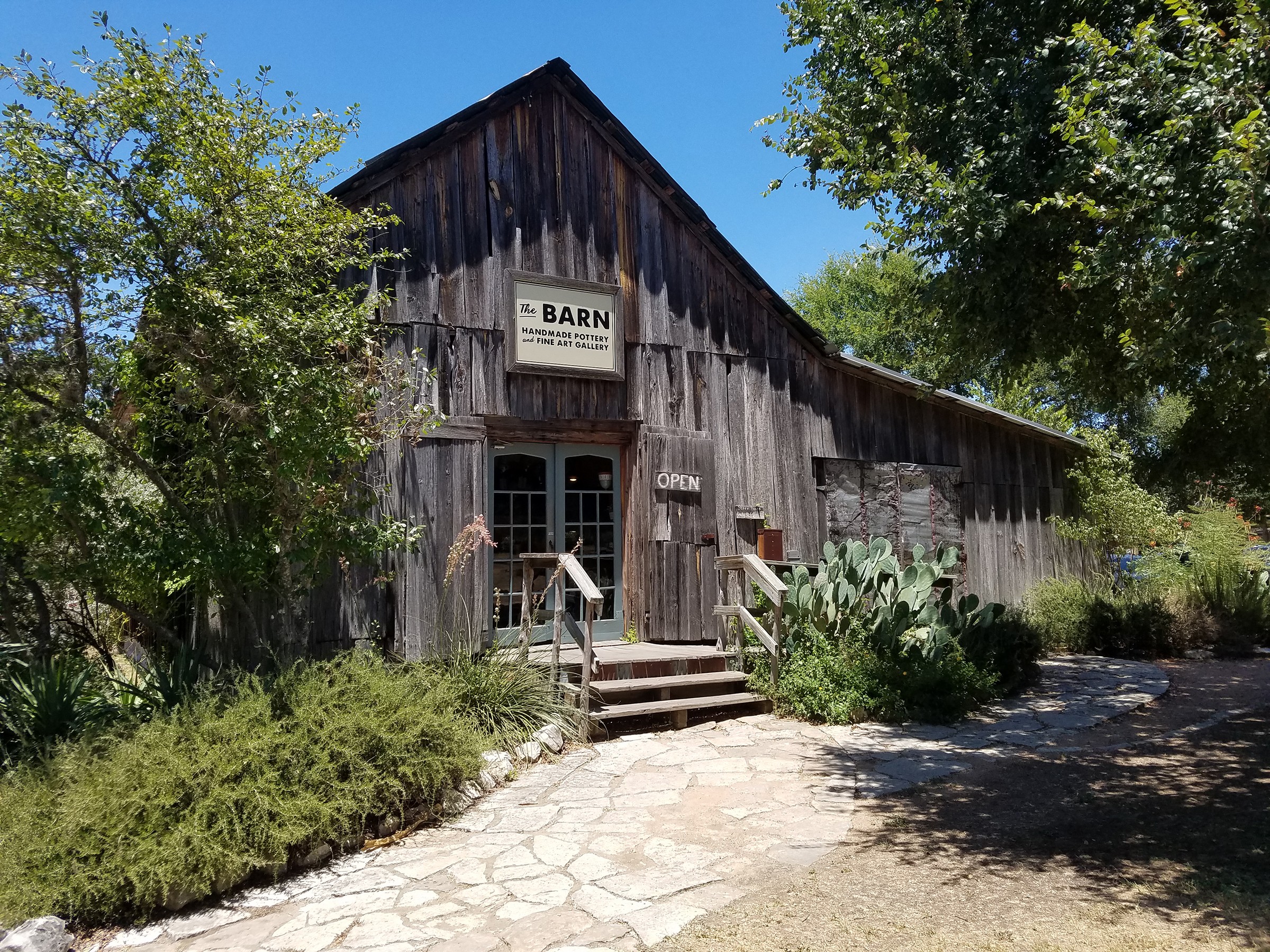 Old wooden barn with a sign, surrounded by trees and plants, under a clear blue sky.