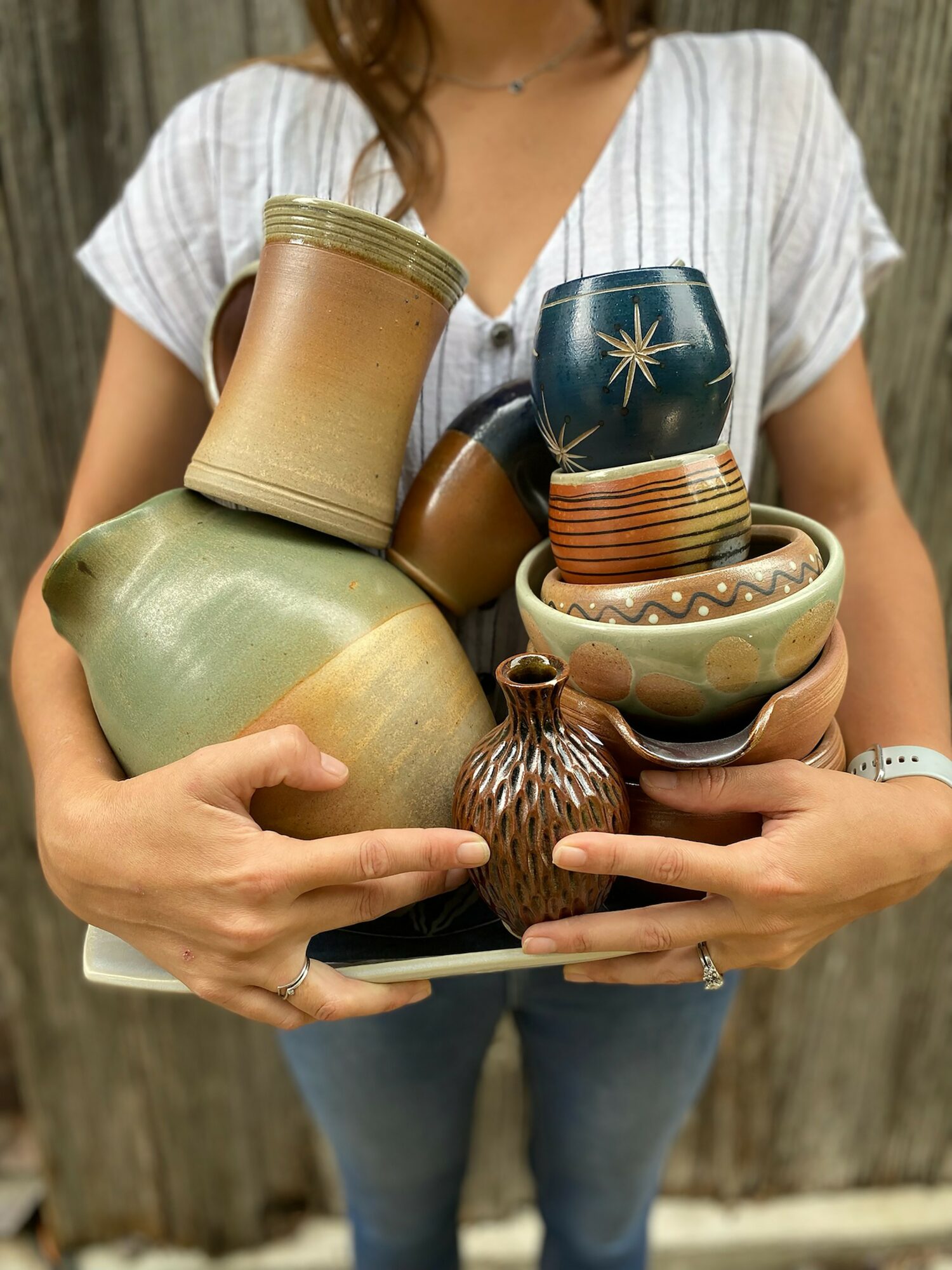 Person holding a tray with various pottery bowls and vases, wearing a white top and jeans.